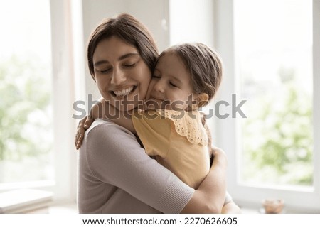 Cheerful happy young mother holding daughter kid in arms, tightening with cheek touches and closed eyes, hugging girl with love, tenderness, devotion, enjoying motherhood, closeness