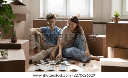 Happy young couple of new homeowners choosing samples for decoration of new apartment, flat, sitting on floor, reviewing brochure with colorful swatches for renovation