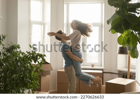 Happy excited young couple celebrating relocation, buying flat, apartment, moving in together, dancing, having fun, laughing. Guy holding girlfriend in arms, spinning at cardboard boxes
