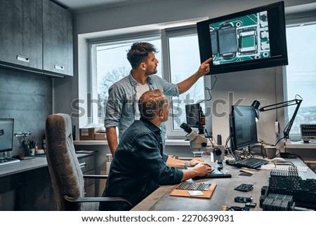 Work in the office. Two men work in a modern office on the development of microchips for the maintenance of electronic devices. The man points to the image of the chip displayed on the monitor.
