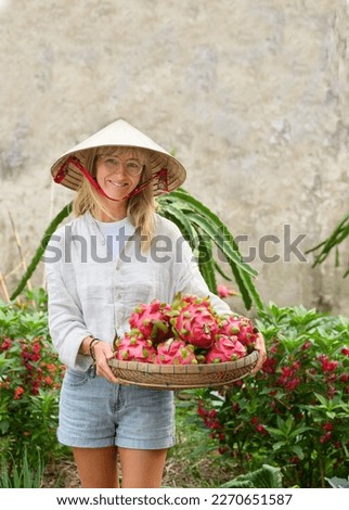 Positive adult female in white shirt and conical hat standing in garden and carrying wicker basket with dragon fruits Royalty-Free Stock Photo #2270651587