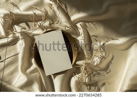 Aesthetic luxury bohemian branding or invitation card template. Blank paper card sheet with empty mock up copy space, dried grass stems on crumpled glossy gold silk background with sunlight shadows