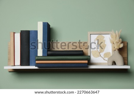Wooden shelf with many hardcover books, picture and dry flowers on light green wall