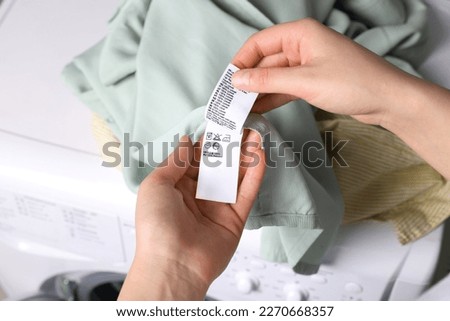Woman reading clothing label with care symbols and material content on green shirt near washing machine, closeup