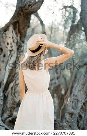 Girl stands in front of a tree holding a straw hat with her hand. Back view