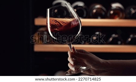 Close up female hand swirling red wine in wine glass. Wine expert tasting, rating and drinking wine, bottles in background. Royalty-Free Stock Photo #2270714825