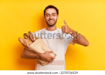 Cheerful caucasian man baker keeps thumb raised shows his agreement holds bunch of fresh crispy baguettes covered with paper smeared with flour wears white t-shirt and apron isolated over yellow