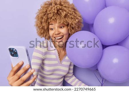 Happy curly haired woman takes selfie via smartphone holds bunch of inflated balloons celebrates special occasion wears striped jumper isolated over purple background. People and celebration concept