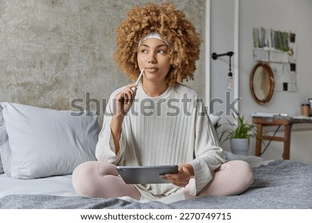 Thoughtful curly haired young woman dressed in nightwear sits crossed legs holds tablet and stylus creats project poses on comfortable bed in bedroom works freelance thinks about future work.