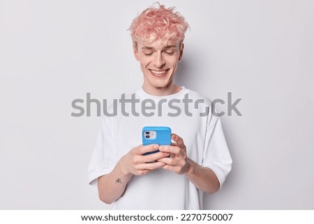 Photo of cheerful man with dyed pink hair laughs at meme his friend sent to him holds mobile phone in hands asnwers on received message dressed in casual t shirt isolated over white background.