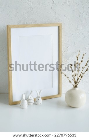 Easter background. Wooden frame mockup with copy space,spring willow branches in a vase and Easter eggs and funny bunny decor near white textured wall in neutral colors.sstkEaster