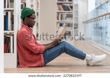 Focused Black student man book lover spending free leisure time in library, African American guy with book in hands sits on floor near bookcase enjoying reading. Hobby, education, university concept Royalty-Free Stock Photo #2270832197