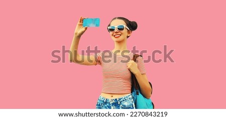 Portrait of happy smiling young woman taking selfie with smartphone wearing backpack on pink background