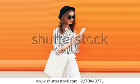 Portrait of beautiful young woman looking at smartphone with shopping bag wearing white striped shirt, black round hat on orange background Royalty-Free Stock Photo #2270843771