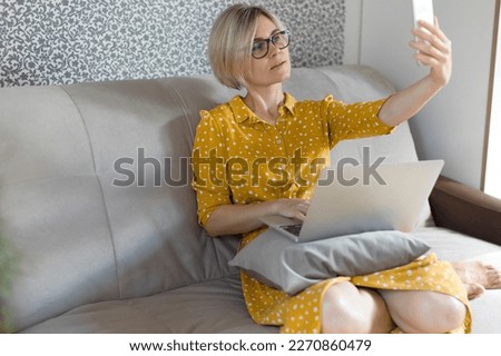 The girl takes a selfie at home against the background of the wall. A blonde woman in an orange dress with a laptop sits on the couch and takes her selfie.