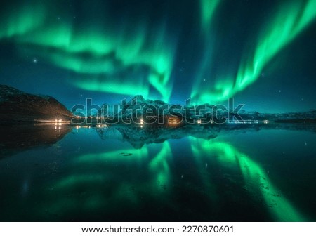 Northern lights over the snowy mountains, sea, reflection in water at night in Lofoten, Norway. Aurora borealis and snow covered rocks. Winter landscape with polar lights, city lights, sky with stars