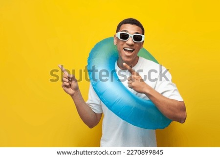 african american guy in sunglasses with inflatable swim ring shows his hands to side on yellow isolated background, man in summer on beach advertises copy space, travel and summer vacation concept Royalty-Free Stock Photo #2270899845