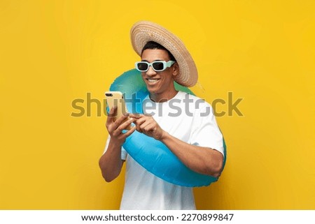 cheerful young guy afro american with inflatable swim ring uses smartphone on yellow isolated background, man in summer on beach is typing on phone, concept of travel and summer holidays