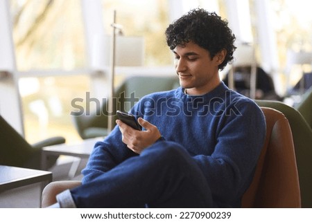 Relaxed young Latin American man using mobile phone, checking social media content, scrolling news feed, messaging, posting photos on his social account, ordering food, booking. People and technology Royalty-Free Stock Photo #2270900239