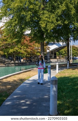 an African American woman with long sisterlocks wearing white and pink clothes, sunglasses and an orange head scarf holding an IPhone walking along a footpath near a pool surrounded by autumn trees	