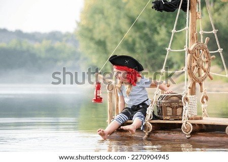 Child in a pirate costume plays on a wooden raft at sunset. Girl pretends to be the captain of a ship with black sails and a flag. Funny kid dreams of adventure and travel.