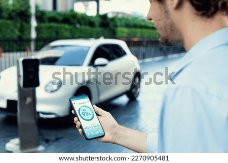 Closeup progressive businessman look at EV car's battery status application on smart phone screen at public parking charging station with power cable plug and renewable energy-powered electric vehicle