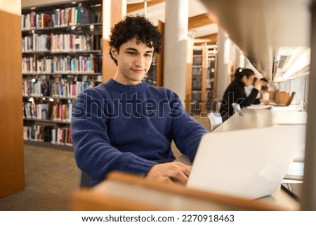 Latin American male student using laptop, focused on working on a diploma project, making researches on internet, studying online in a modern innovation library campus. People. Erudition. Education.