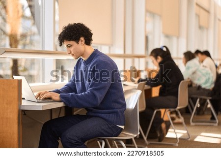 Multitasking, overworked, serious Latin American Young male student, online studying in a modern innovation library campus, sitting at a desk and typing text on laptop keyboard, searching info on net Royalty-Free Stock Photo #2270918505