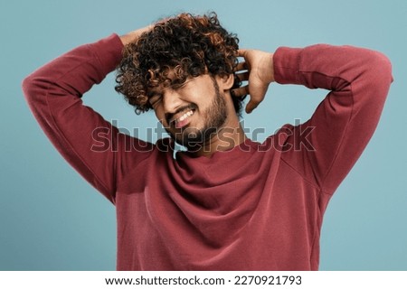 Young curly-haired bearded Indian man scratching his head on isolated blue background. Pediculosis. Annoying itch. Man with lice and nits. Concept with Healthcare And Medicine.
