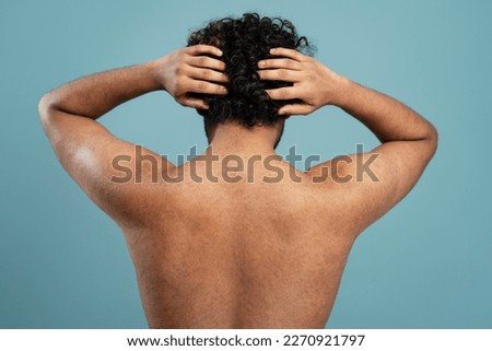 Young shirtless dark-haired curly man scratching head or massaging head skin standing back to the camera, isolated on blue background. Skin, body and hair care concept. Male beauty portrait