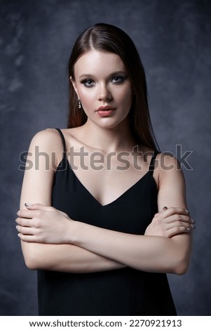 Portrait of female in black tank top with straps. Woman with earrings in the ear on gray background. Hands crossed.