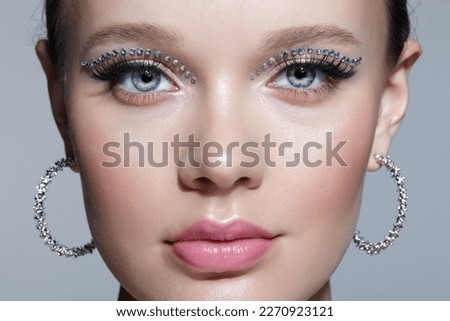 Closeup shot of human female face with unusual rhinestones makeup. Woman with earring in the form of a shiny ring in the ear on gray background. Royalty-Free Stock Photo #2270923121
