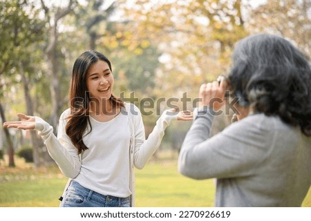 Happy Asian grandmother taking a picture of her lovely granddaughter with a camera while strolling in the green park together. family bonding