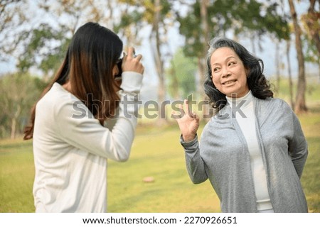 Lovely Asian granddaughter taking a picture of her happy grandmother with retro camera while spending time together in the beautiful green park. leisure concept