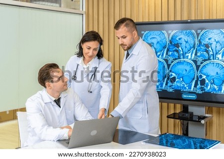 Team of three doctors discusses the results of an MRI scan of the patient's head in the consulting room of modern hospital, physician discussing about treatment for patient looking in laptop Royalty-Free Stock Photo #2270938023