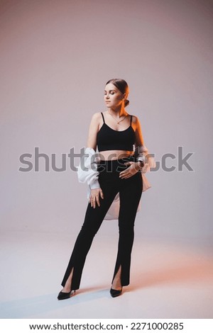 A young slender woman in a white shirt and black pants with makeup on her face poses on a plain background. Stylish European woman in a black top.