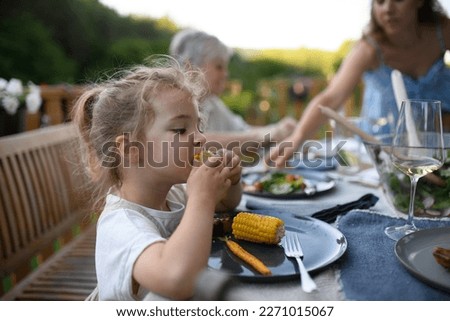 Family eating at barbecue party dinner on patio, little girl eating roasted corn and enjoying it.