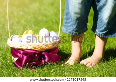 A child stands with bare feet on a green lawn, Easter eggs basket. Beautiful spring photo with Easter eggs in a wicker basket decorated with a purple bow. The concept of celebrating Easter