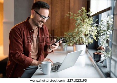 Satisfied modern millennial man in stylish casual clothes using smartphone and laptop computer for electronic banking, making reservation, online shopping and payments while spending time at home. Royalty-Free Stock Photo #2271023195
