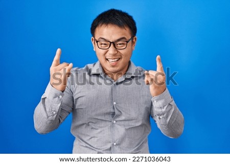Young chinese man standing over blue background shouting with crazy expression doing rock symbol with hands up. music star. heavy concept. 
