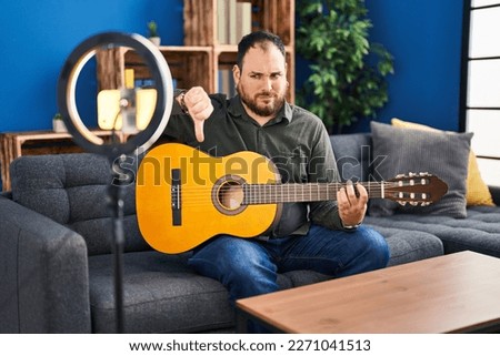 Plus size hispanic man with beard playing classic guitar at music studio recording himself with angry face, negative sign showing dislike with thumbs down, rejection concept 