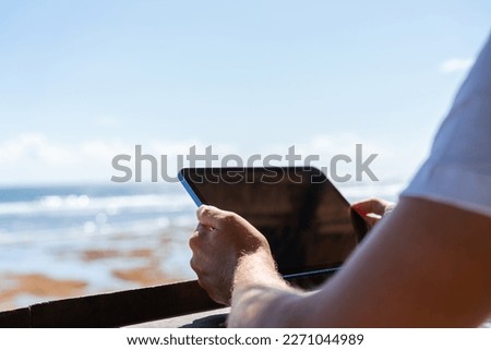 Young man hands holding a tablet, blurred background of beach and empty copy space sky. Online connection and internet browse. Concept of journey and tourism