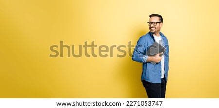 Smiling man holding laptop and looking over the shoulder. Cheerful handsome guy standing against yellow background. He is wearing casual outfit