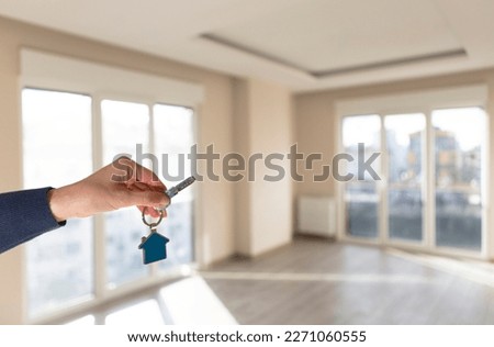Real estate, house rental, housing sale and mortgage concepts. Woman's hand holding new house key, empty living room of apartment for sale or rent in background. Horizontal close up.