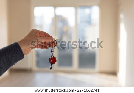 Woman's hand holding new house key, empty living room of apartment for sale or rent in background. Real estate, house rental, housing sale and mortgage concepts. Horizontal close up.
