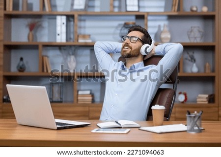 Mature businessman in blue shirt relaxing in office, man with headphones folded hands on title, listening to soothing music and audiobook podcasts, boss at workplace sitting using laptop.