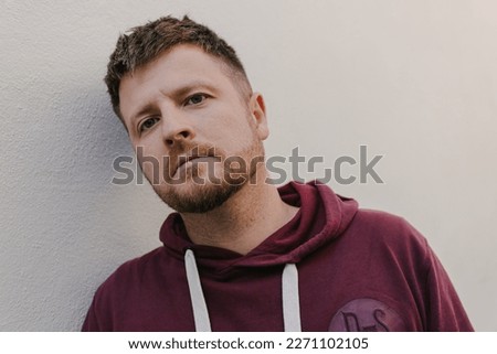 Man is standing near a white wall. He is wearing a burgundy hoodie. Close-up portrait. 30-35 year old European man with crop haircut. He is fair-haired with beard and grey eyes. Royalty-Free Stock Photo #2271102105