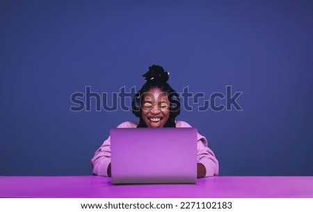 Cheerful female gamer smiling excitedly while playing a video game on a laptop. Happy young woman winning an online game in a studio. Young black woman streaming a game against a purple background.