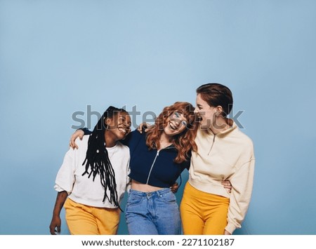 Multicultural female friends laughing and having fun while embracing each other. Group of cheerful young women enjoying themselves while standing against a blue background. Royalty-Free Stock Photo #2271102187