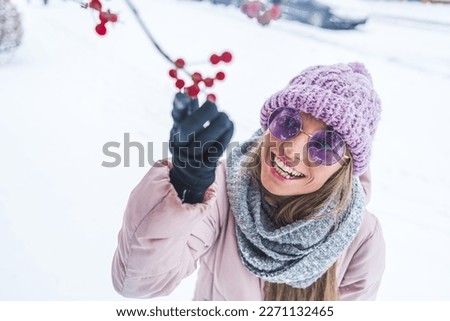 cheerful woman wearing a coat, a scarf, a hat and gloves touching tree red fruit in the snow, medium closeup . High quality photo